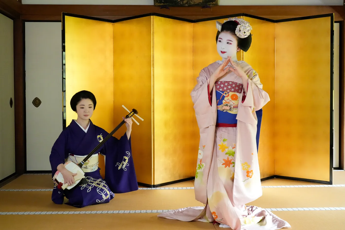 A maiko dancing in front of a gold folding screen, accompanied by a geiko on the shamisen