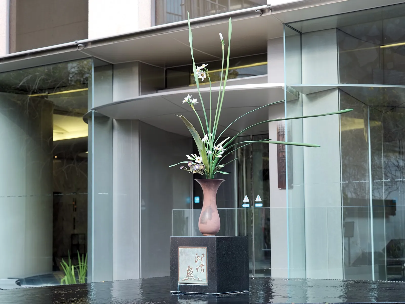 A tall ikebana arrangement in a vase, displayed in a glass case at the foot of the Ikenobo building beside Rokkakudo