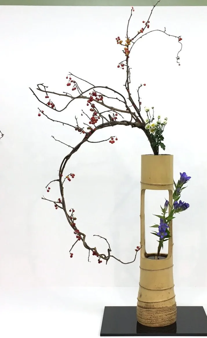 An ikebana arrangement with a branch of red berries curving in a wide arc above a tall bamboo vessel, with blue flowers rising through an opening in the bamboo