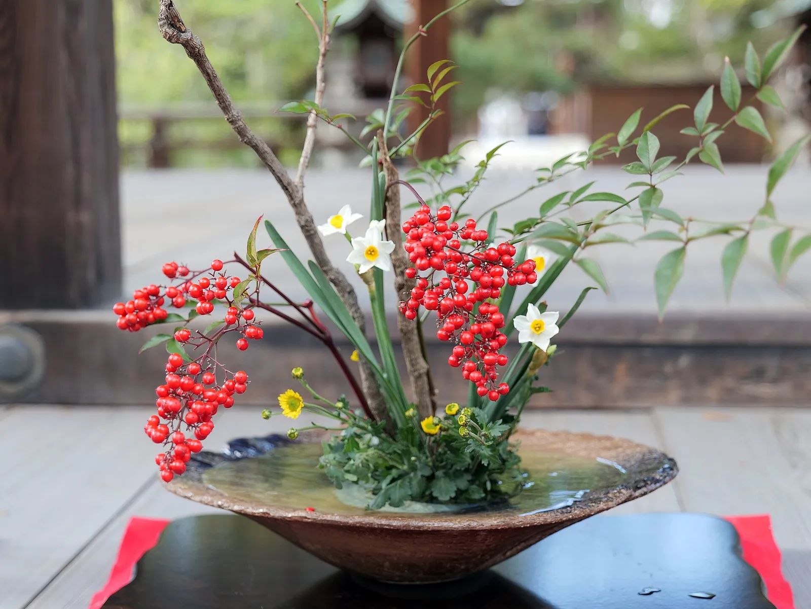 An ikebana arrangement of red berries, white and yellow flowers, and long green leaves in a wide, shallow bowl on the wooden floor of a shrine