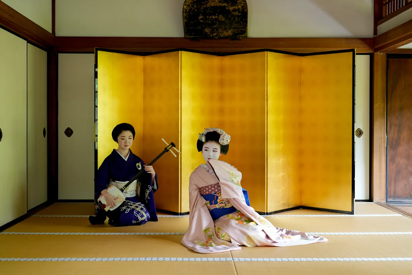 A maiko bowing on tatami before a gold folding screen at the end of a dance, with a geiko holding a shamisen