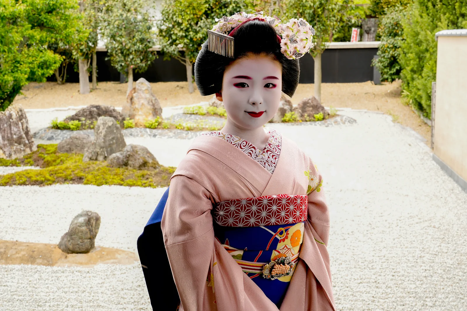 A maiko with hair ornaments and white makeup in a rock garden