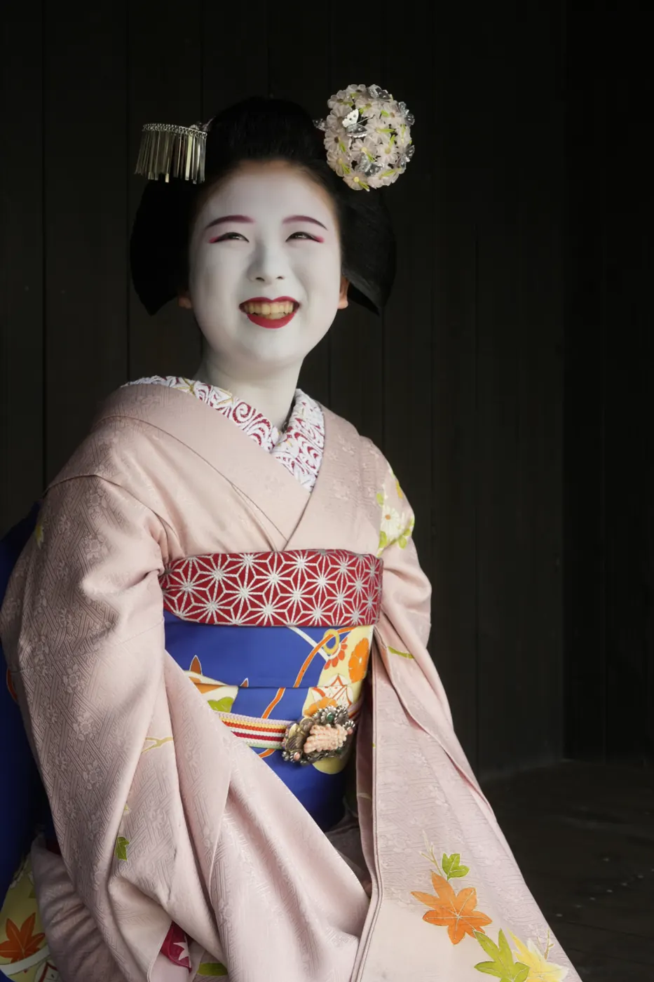 A smiling maiko in white makeup with a flower hairpin and a silver hair ornament in her own hair