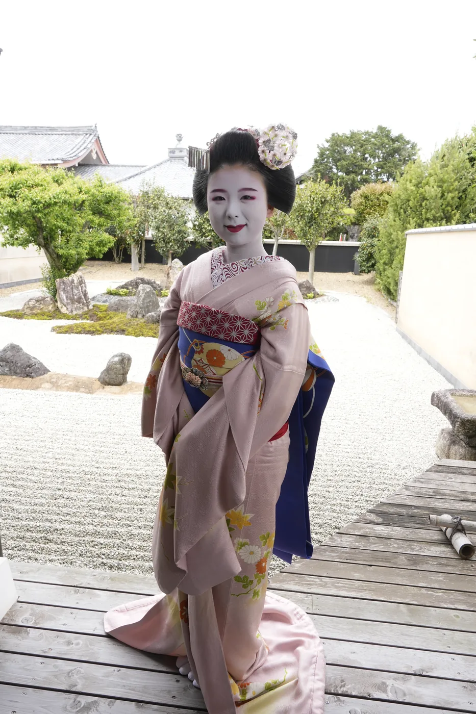 A maiko in a pink kimono with seasonal motifs standing on a wooden veranda beside a raked gravel garden