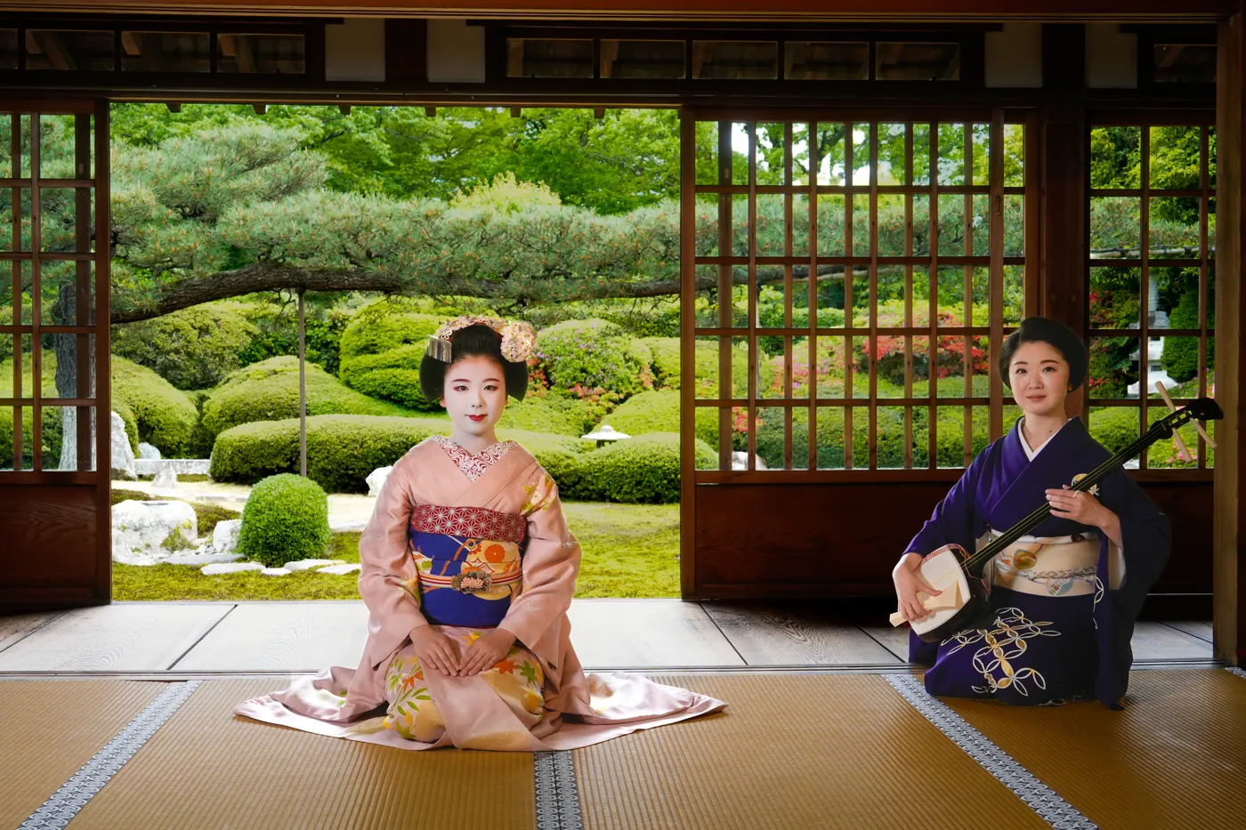 A maiko and a geiko shamisen player kneeling on tatami with a temple garden behind them