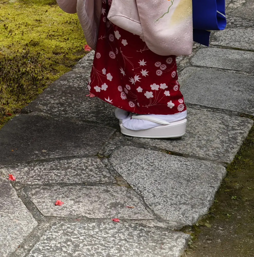 Close view of a maiko's red patterned underlayer and tall white okobo sandals on a stone path