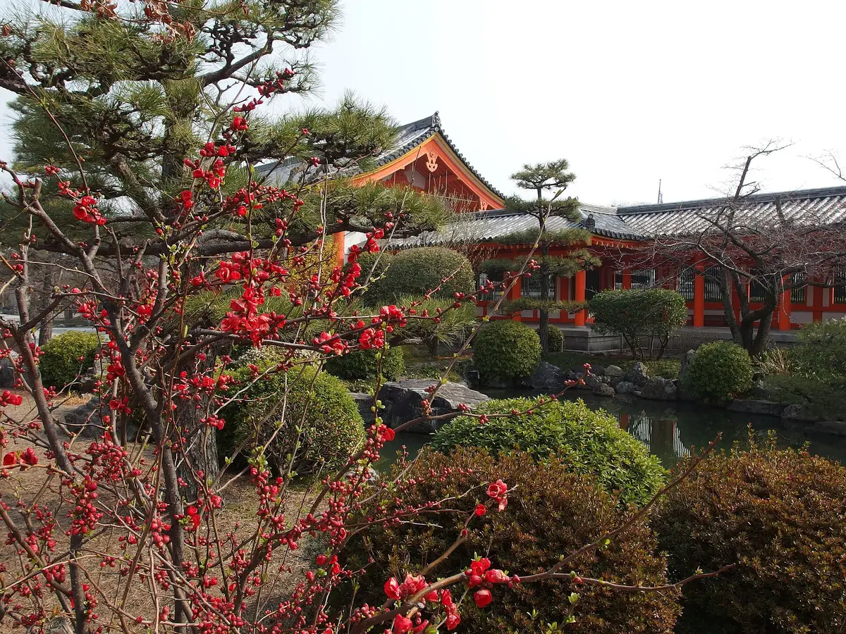 A pond garden at Sanjusangendo with red flowering branches and an orange-red building behind