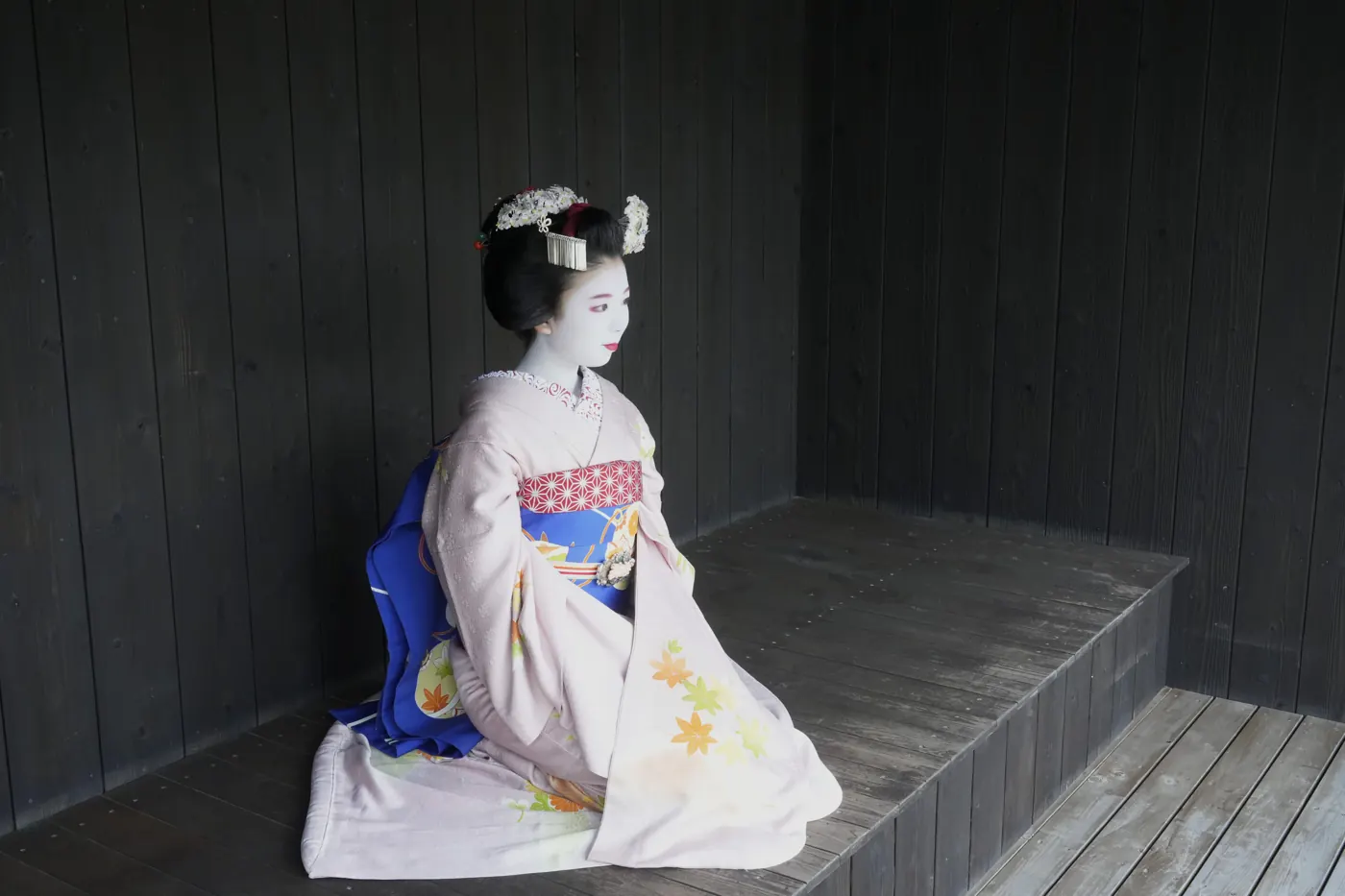 A maiko kneeling on a wooden veranda in a pale kimono with a blue obi